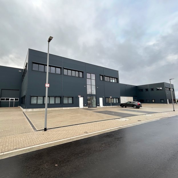 Moderne, dunkelgraue Industriehalle mit großen Fenstern und einer zentralen Glasfassade als Eingangsbereich. Die Halle steht auf einem gepflasterten Gelände mit mehreren Parkplätzen, von denen einer mit einem schwarzen Auto belegt ist. Eine Straßenlaterne mit einem kleinen roten Schild steht im Vordergrund. Der Himmel ist bewölkt, und die Straße vor der Halle ist nass.