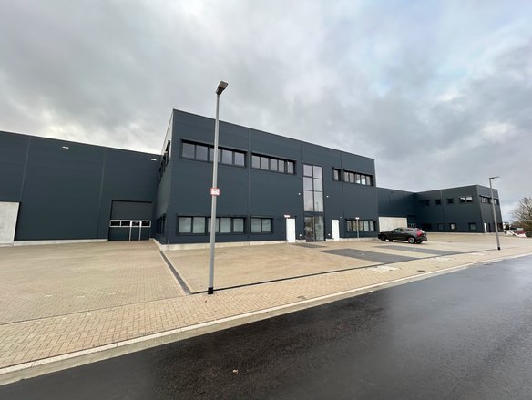 Moderne, dunkelgraue Industriehalle mit großen Fenstern und einer zentralen Glasfassade als Eingangsbereich. Die Halle steht auf einem gepflasterten Gelände mit mehreren Parkplätzen, von denen einer mit einem schwarzen Auto belegt ist. Eine Straßenlaterne mit einem kleinen roten Schild steht im Vordergrund. Der Himmel ist bewölkt, und die Straße vor der Halle ist nass.