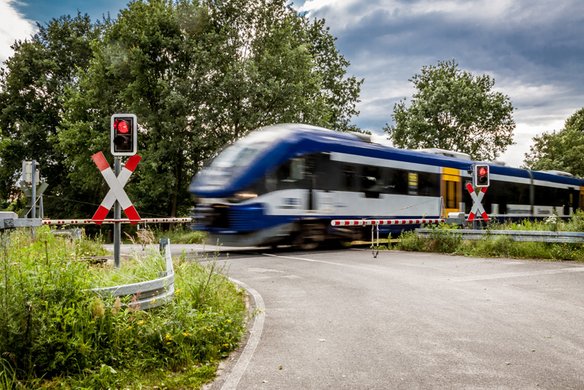 Ein Regionalzug mit blau-weißer Lackierung fährt mit hoher Geschwindigkeit über einen Bahnübergang. Die Schranken sind geschlossen, und die roten Warnlichter leuchten. Links und rechts stehen Andreaskreuze zur Kennzeichnung des Bahnübergangs. Die Straße ist von grüner Vegetation umgeben, und im Hintergrund sind Bäume sowie ein bewölkter Himmel zu sehen.