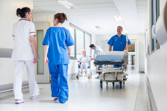Krankenhausflur mit medizinischem Personal: Zwei Pflegekräfte in blauer und weißer Arbeitskleidung gehen den Gang entlang, während ein Arzt in blauer Kleidung ein leeres Krankenhausbett schiebt. Im Hintergrund sitzt eine ältere Patientin im Rollstuhl, betreut von einem weiteren Arzt.