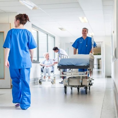 Krankenhausflur mit medizinischem Personal: Zwei Pflegekräfte in blauer und weißer Arbeitskleidung gehen den Gang entlang, während ein Arzt in blauer Kleidung ein leeres Krankenhausbett schiebt. Im Hintergrund sitzt eine ältere Patientin im Rollstuhl, betreut von einem weiteren Arzt.