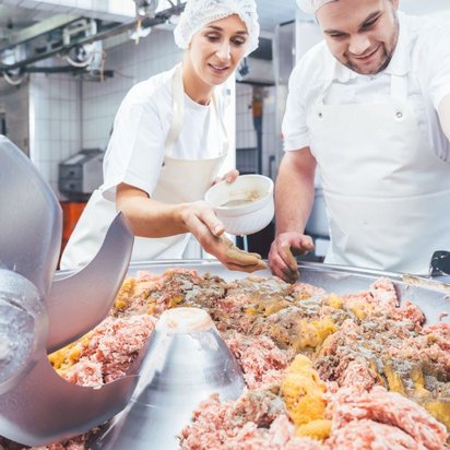 Zwei Metzger oder Lebensmittelverarbeiter, eine Frau und ein Mann, arbeiten in einer Fleischerei oder Lebensmittelproduktion. Sie tragen weiße Arbeitskleidung, Schürzen und Haarnetze. Die Frau streut mit der Hand Gewürze aus einer Schale auf eine große Menge Hackfleisch in einer industriellen Kuttermaschine. Der Mann verteilt die Gewürze weiter auf der Fleischmasse. Im Vordergrund sind die scharfen Kuttermesser sichtbar, während der Hintergrund eine saubere Produktionshalle mit Fliesenwänden und Edelstahlgeräten zeigt.