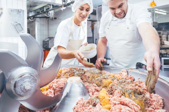 Zwei Metzger oder Lebensmittelverarbeiter, eine Frau und ein Mann, arbeiten in einer Fleischerei oder Lebensmittelproduktion. Sie tragen weiße Arbeitskleidung, Schürzen und Haarnetze. Die Frau streut mit der Hand Gewürze aus einer Schale auf eine große Menge Hackfleisch in einer industriellen Kuttermaschine. Der Mann verteilt die Gewürze weiter auf der Fleischmasse. Im Vordergrund sind die scharfen Kuttermesser sichtbar, während der Hintergrund eine saubere Produktionshalle mit Fliesenwänden und Edelstahlgeräten zeigt.