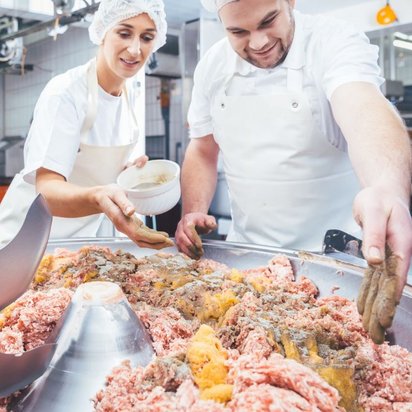 Zwei Metzger oder Lebensmittelverarbeiter, eine Frau und ein Mann, arbeiten in einer Fleischerei oder Lebensmittelproduktion. Sie tragen weiße Arbeitskleidung, Schürzen und Haarnetze. Die Frau streut mit der Hand Gewürze aus einer Schale auf eine große Menge Hackfleisch in einer industriellen Kuttermaschine. Der Mann verteilt die Gewürze weiter auf der Fleischmasse. Im Vordergrund sind die scharfen Kuttermesser sichtbar, während der Hintergrund eine saubere Produktionshalle mit Fliesenwänden und Edelstahlgeräten zeigt.