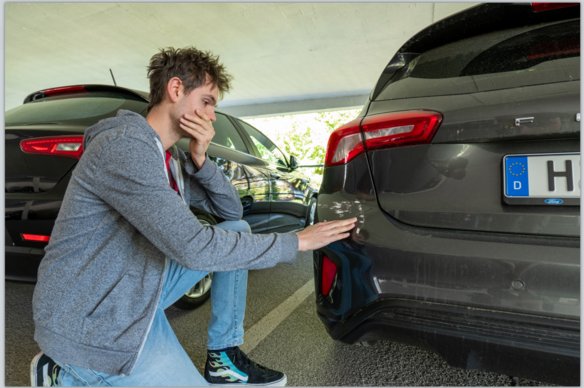 Ein junger Mann mit zerzausten Haaren, grauer Kapuzenjacke und blauen Jeans kniet in einem überdachten Parkplatz neben einem Auto. Er hält eine Hand nachdenklich an seinen Mund und berührt mit der anderen Hand einen Kratzer an der hinteren Stoßstange eines dunklen Fahrzeugs. Im Hintergrund sind weitere parkende Autos zu sehen.