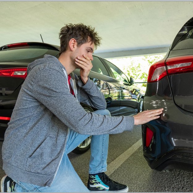 Ein junger Mann mit zerzausten Haaren, grauer Kapuzenjacke und blauen Jeans kniet in einem überdachten Parkplatz neben einem Auto. Er hält eine Hand nachdenklich an seinen Mund und berührt mit der anderen Hand einen Kratzer an der hinteren Stoßstange eines dunklen Fahrzeugs. Im Hintergrund sind weitere parkende Autos zu sehen.