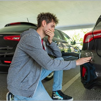 Ein junger Mann mit zerzausten Haaren, grauer Kapuzenjacke und blauen Jeans kniet in einem überdachten Parkplatz neben einem Auto. Er hält eine Hand nachdenklich an seinen Mund und berührt mit der anderen Hand einen Kratzer an der hinteren Stoßstange eines dunklen Fahrzeugs. Im Hintergrund sind weitere parkende Autos zu sehen.