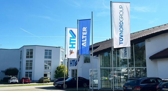 Drei Fahnenmasten vor einem modernen Bürogebäude mit Glasfassade. Die linke Fahne zeigt das HTV-Logo, die mittlere das Wort "ALTER" in blauen Buchstaben, die rechte das TÜV NORD GROUP-Logo