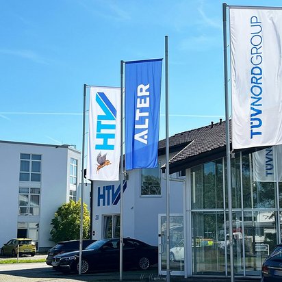 Drei Fahnenmasten vor einem modernen Bürogebäude mit Glasfassade. Die linke Fahne zeigt das HTV-Logo, die mittlere das Wort "ALTER" in blauen Buchstaben, die rechte das TÜV NORD GROUP-Logo