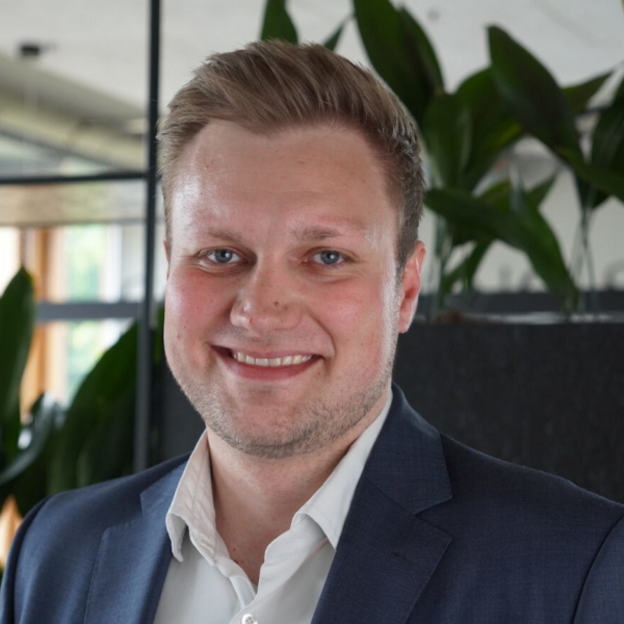 Ein junger Mann mit kurzen, blond-braunen Haaren lächelt in die Kamera. Er trägt einen dunkelblauen Anzug mit einem weißen Hemd. Im Hintergrund befinden sich grüne Pflanzen und moderne Büroelemente
