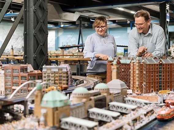 A man and a woman admire a detailed miniature city model featuring historic buildings, bridges, and waterways in an indoor exhibition setting