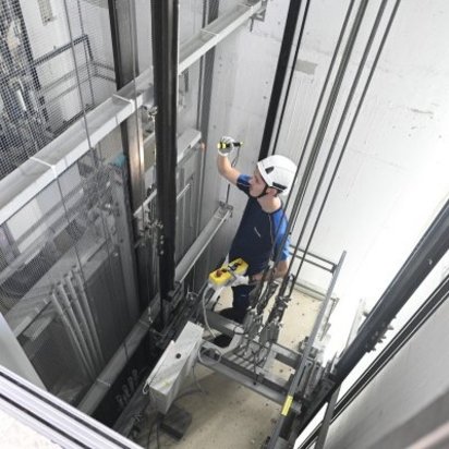 Ein Techniker mit Schutzhelm und blauer Arbeitskleidung steht im Fahrstuhlschacht und führt eine Inspektion durch. Er hält eine Taschenlampe in der Hand und überprüft die Aufzugsmechanik und Seilzüge. Die Umgebung ist von Metallgittern und technischen Komponenten umgeben.