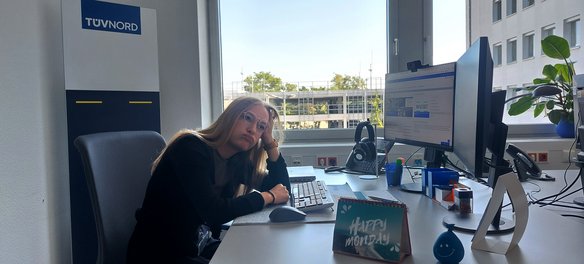 Eine junge Frau mit Brille und langen blonden Haaren sitzt an einem Schreibtisch in einem Büro mit TÜV NORD-Branding. Sie stützt ihren Kopf auf die Hand und blickt müde oder gelangweilt. Vor ihr stehen zwei Monitore, eine Tastatur, ein Telefon und verschiedene Büroutensilien. Ein kleines Aufstell-Schild auf dem Tisch trägt die Aufschrift „Happy Monday“. Durch das Fenster ist ein Bürogebäude und etwas Grün zu sehen.