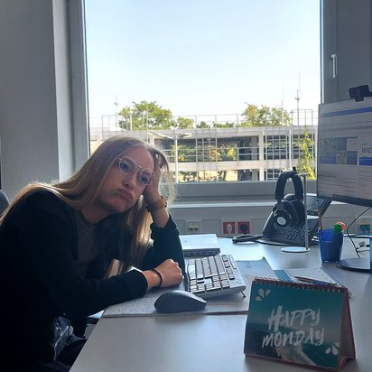 Eine junge Frau mit Brille und langen blonden Haaren sitzt an einem Schreibtisch in einem Büro mit TÜV NORD-Branding. Sie stützt ihren Kopf auf die Hand und blickt müde oder gelangweilt. Vor ihr stehen zwei Monitore, eine Tastatur, ein Telefon und verschiedene Büroutensilien. Ein kleines Aufstell-Schild auf dem Tisch trägt die Aufschrift „Happy Monday“. Durch das Fenster ist ein Bürogebäude und etwas Grün zu sehen.