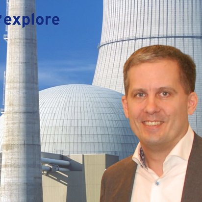Mann im Anzug mit kurzem, hellbraunem Haar lächelt in die Kamera. Im Hintergrund befindet sich ein großes Kernkraftwerk mit einem hohen Schornstein, einer Kuppel und einem Kühlturm. Oben links im Bild steht der Hashtag „#explore“ in blauer Schrift.