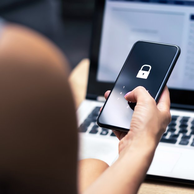 Nahaufnahme einer Hand, die ein Smartphone hält, auf dessen Bildschirm ein Vorhängeschloss-Symbol angezeigt wird. Der Hintergrund ist unscharf und zeigt einen Laptop-Bildschirm mit einer Webseite. Die Hand und das Smartphone sind im Fokus, während der Laptop im Hintergrund verschwimmt