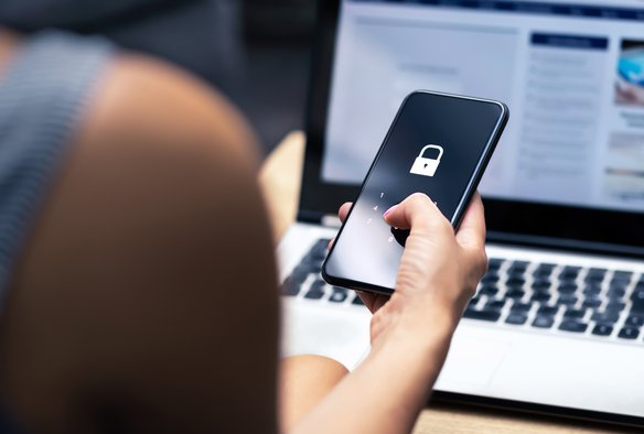 Nahaufnahme einer Hand, die ein Smartphone hält, auf dessen Bildschirm ein Vorhängeschloss-Symbol angezeigt wird. Der Hintergrund ist unscharf und zeigt einen Laptop-Bildschirm mit einer Webseite. Die Hand und das Smartphone sind im Fokus, während der Laptop im Hintergrund verschwimmt