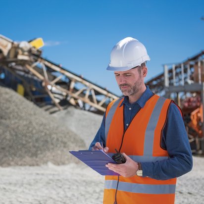 Ein Bauleiter mit weißem Schutzhelm und orangefarbener Warnweste steht auf einer Baustelle mit einer großen Schotterhalde und Förderbändern im Hintergrund. Er hält ein Klemmbrett und ein Funkgerät in den Händen und notiert etwas, während er konzentriert schaut. Der Himmel ist blau und wolkenlos.