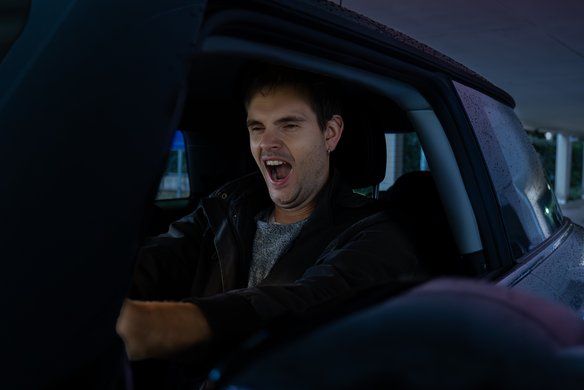Ein junger Mann mit kurzen braunen Haaren und Ohrpiercing sitzt in einem Auto und gähnt mit weit geöffnetem Mund. Er trägt eine dunkle Jacke über einem grauen Pullover. Das Auto befindet sich in einer überdachten Parkstruktur, und durch das Fenster ist eine leicht unscharfe Außenansicht mit Bäumen und Gebäuden zu sehen. Die Beleuchtung wirkt kühl und leicht gedimmt.