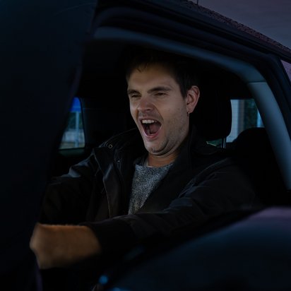 Ein junger Mann mit kurzen braunen Haaren und Ohrpiercing sitzt in einem Auto und gähnt mit weit geöffnetem Mund. Er trägt eine dunkle Jacke über einem grauen Pullover. Das Auto befindet sich in einer überdachten Parkstruktur, und durch das Fenster ist eine leicht unscharfe Außenansicht mit Bäumen und Gebäuden zu sehen. Die Beleuchtung wirkt kühl und leicht gedimmt.