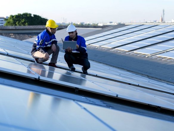 Zwei Ingenieure in blauen Schutzjacken und Sicherheitshelmen knien auf einem großen Dach mit Solarpanels. Einer trägt einen gelben Helm, der andere einen weißen und hält ein Tablet in den Händen. Sie analysieren die Solaranlage bei Tageslicht. Im Hintergrund sind weitere Solarpaneele sowie Bäume und Gebäude sichtbar.