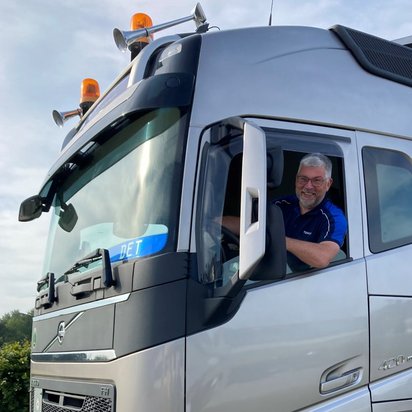 lkw-fahrer-volvo-kabine.jpg Ein lächelnder Lkw-Fahrer mit grauen Haaren und Brille sitzt in der Fahrerkabine eines silbernen Volvo-Lastwagens und schaut aus dem offenen Fenster. Der Lkw ist mit orangefarbenen Rundumleuchten und mehreren Spiegeln ausgestattet.